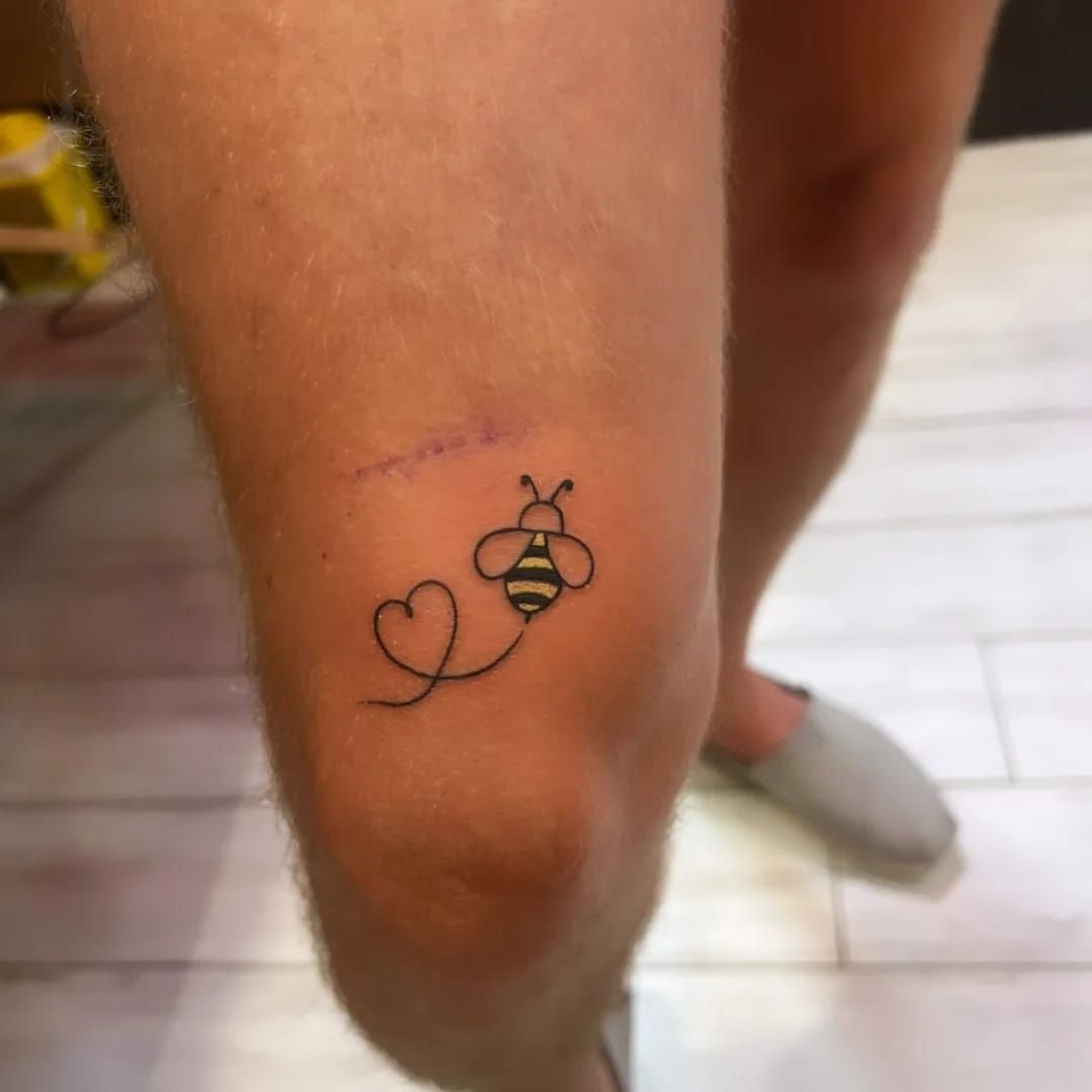 Tatuaje de abeja en la rodilla, con rastro de corazón, tinta negra.
