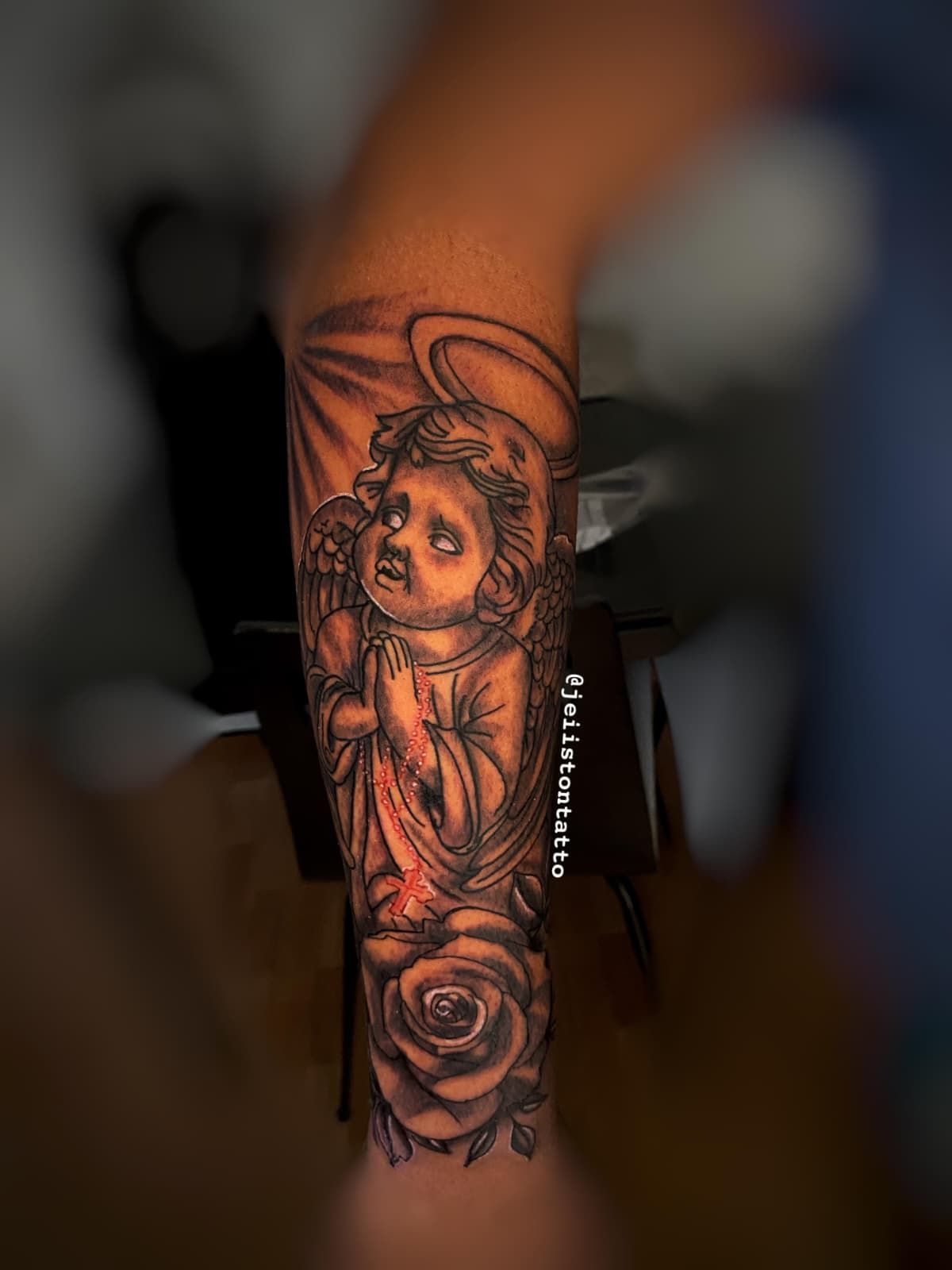 Tatuaje en el brazo: Ángel rezando con halo, una rosa y rayos de sol en escala de grises.