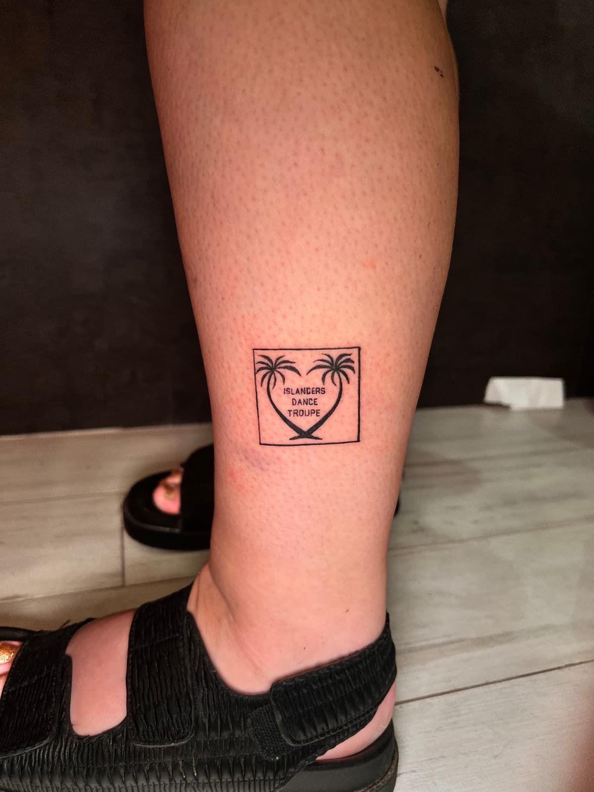 Tatuaje de palmeras dentro de un marco en forma de corazón dentro de un cuadrado, en la pierna de una persona, con sandalias.