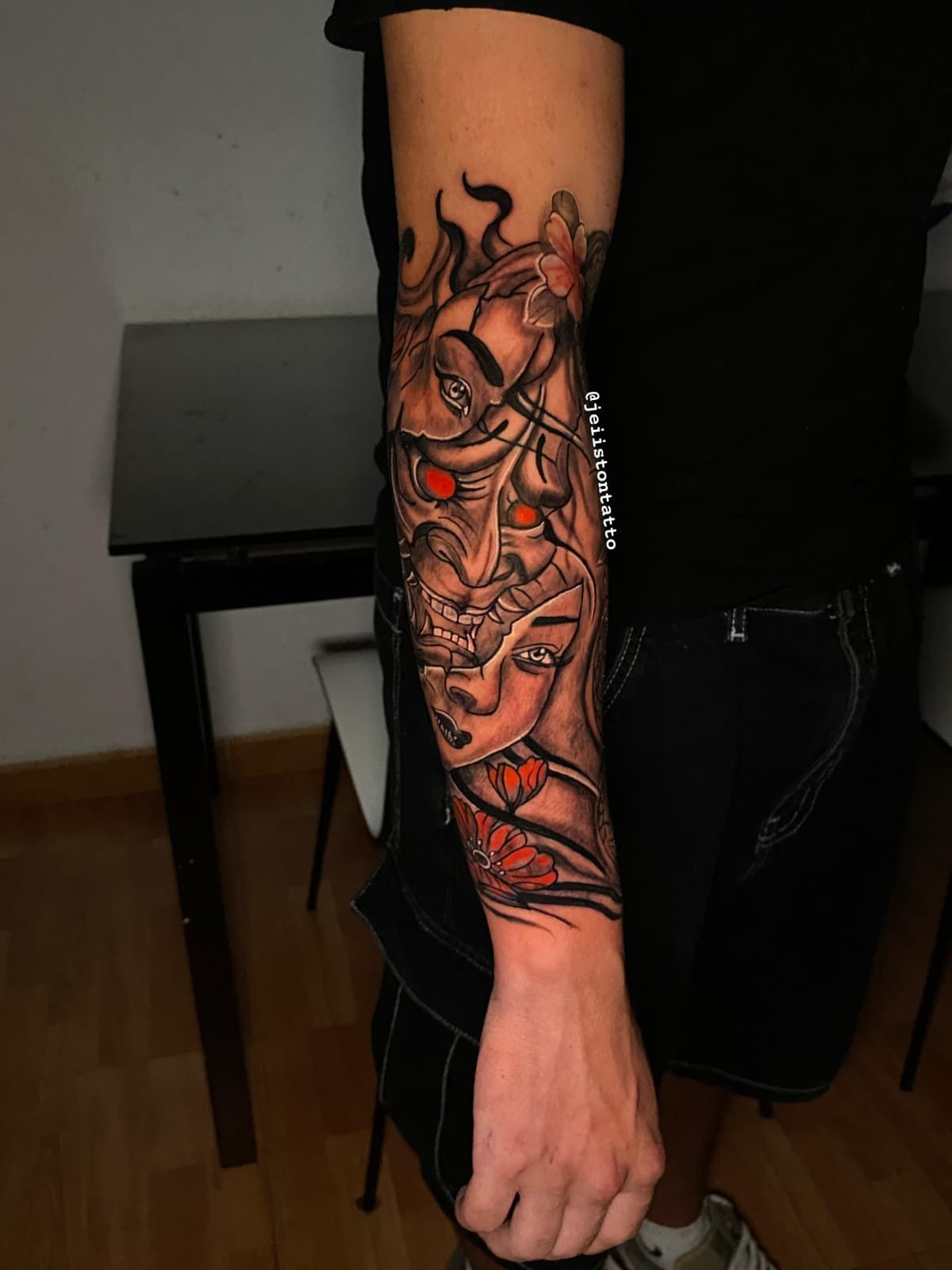 Tatuaje de dos rostros enmascarados en el antebrazo, con acentos ardientes. Ojos rojos y sombreado intrincado.