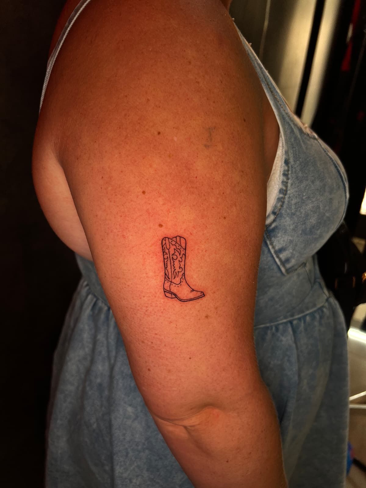 Brazo de mujer con un pequeño tatuaje de bota vaquera negra. Lleva un vestido vaquero.