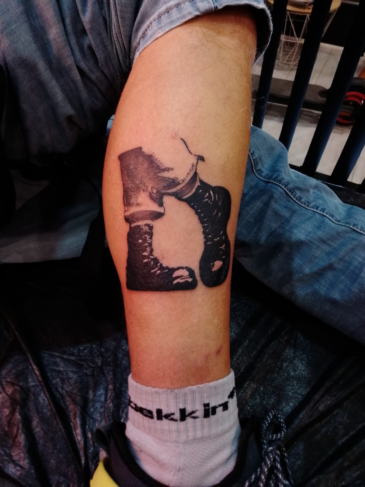 Tatuaje de una bota militar y un calcetín en el antebrazo. La persona lleva vaqueros y calcetín.
