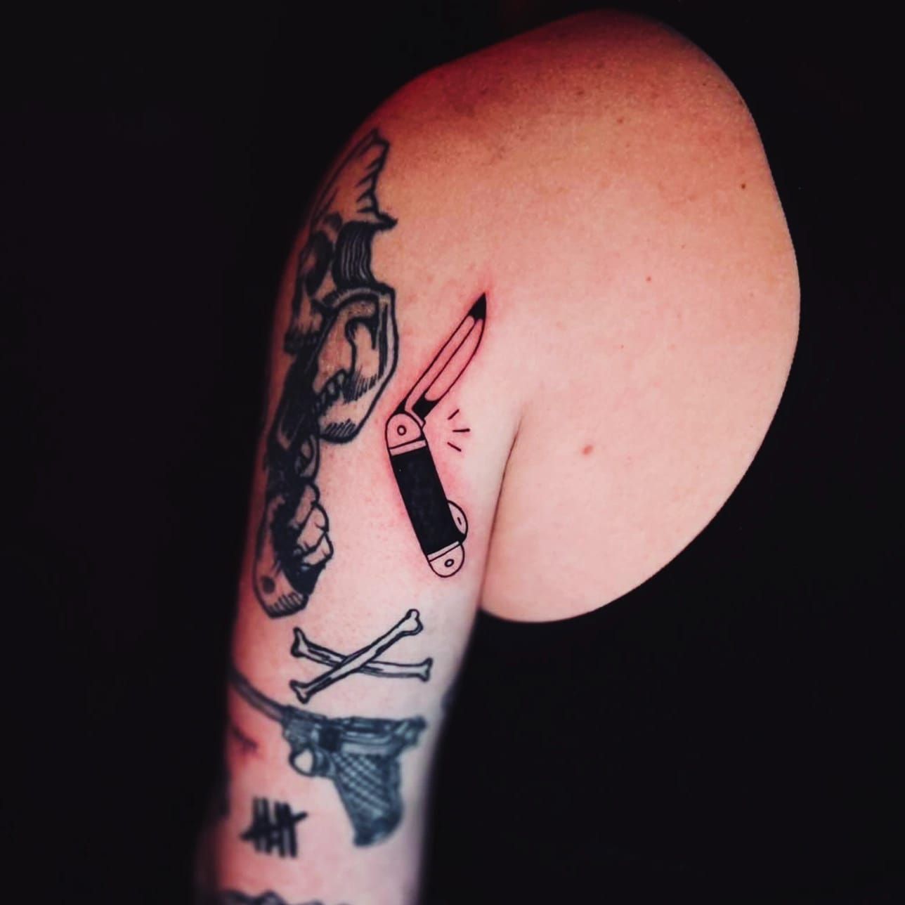 Brazo con tatuajes: calavera, navaja, huesos cruzados, pistola y marcas de conteo.