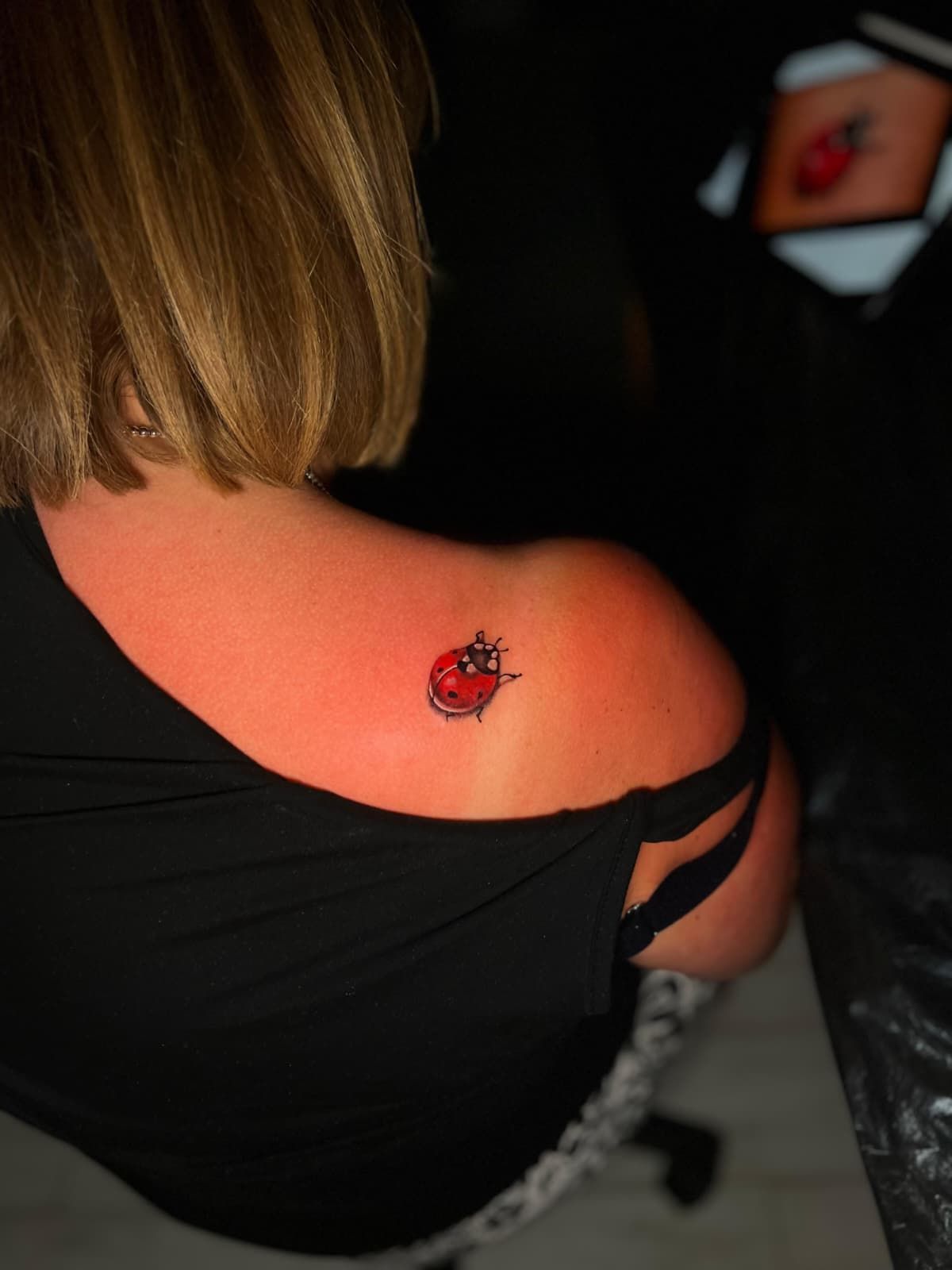 Hombro de mujer con un pequeño tatuaje de mariquita. Top negro, piel clara, quemaduras solares rojas.