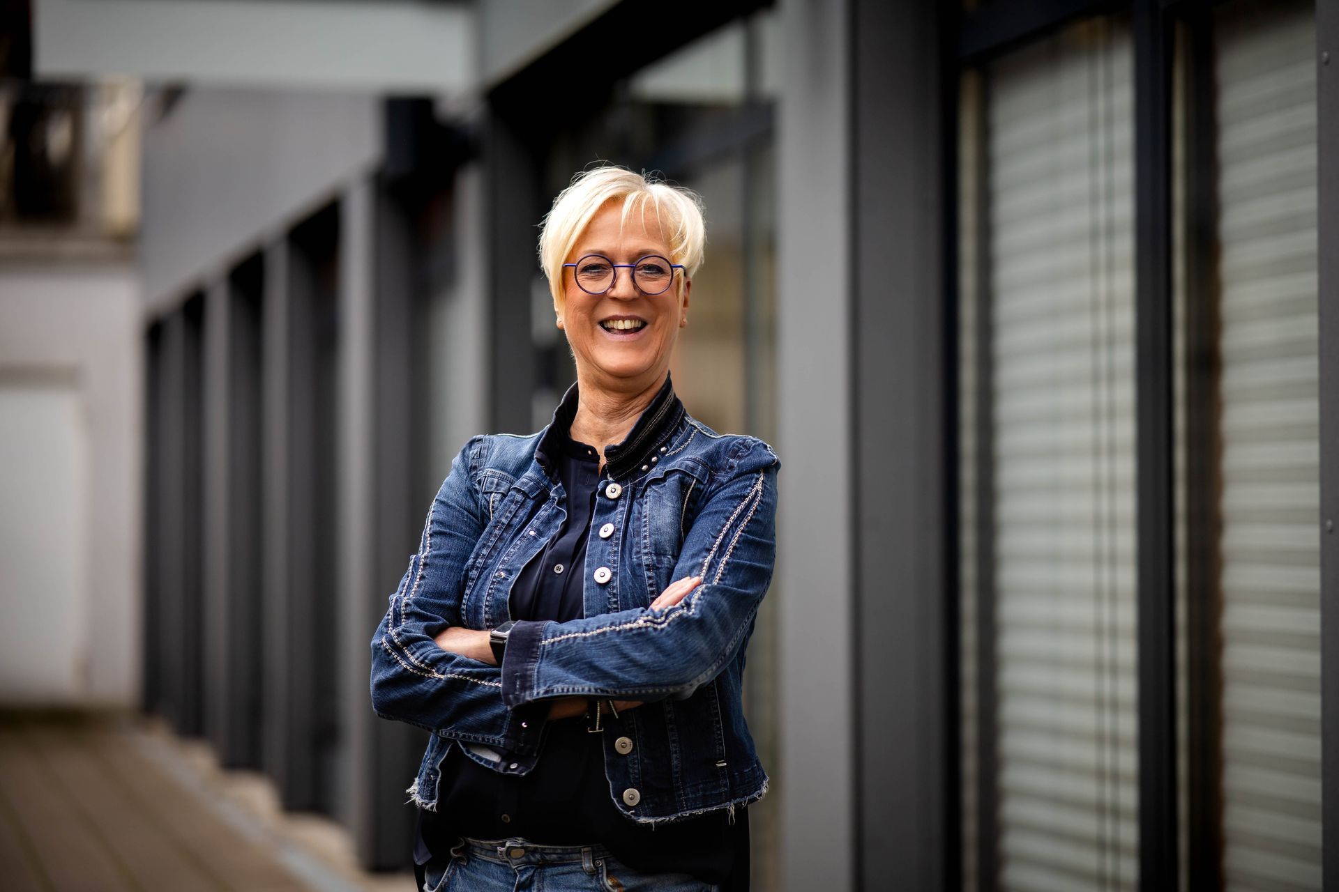 Regina Bicker Portrait Eine Person mit kurzen blonden Haaren und Brille lächelt vor einem hellblauen Hintergrund und trägt eine dunkle Jeansjacke.
