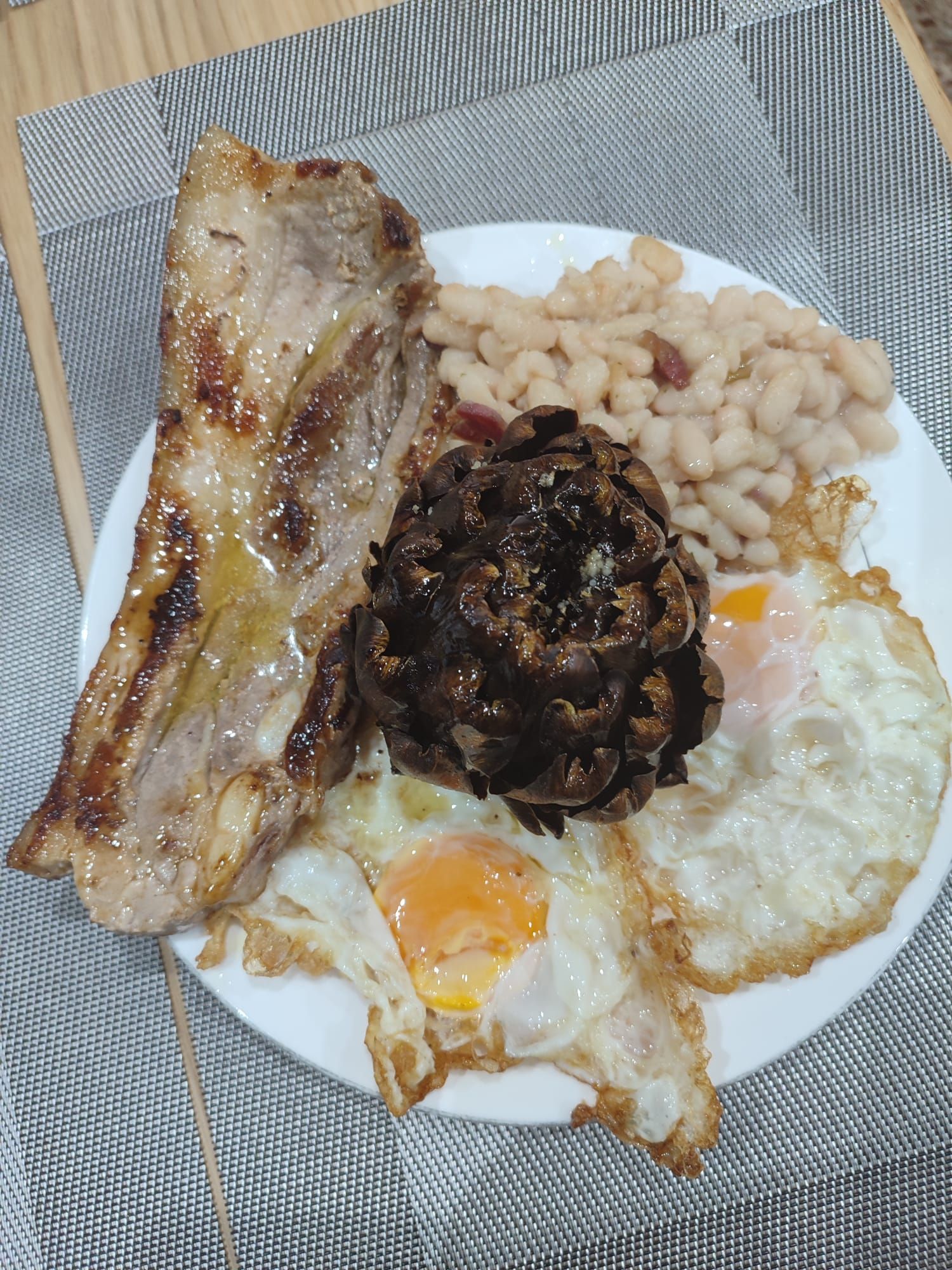 Plato con cerdo frito, frijoles, una alcachofa oscura y dos huevos fritos.