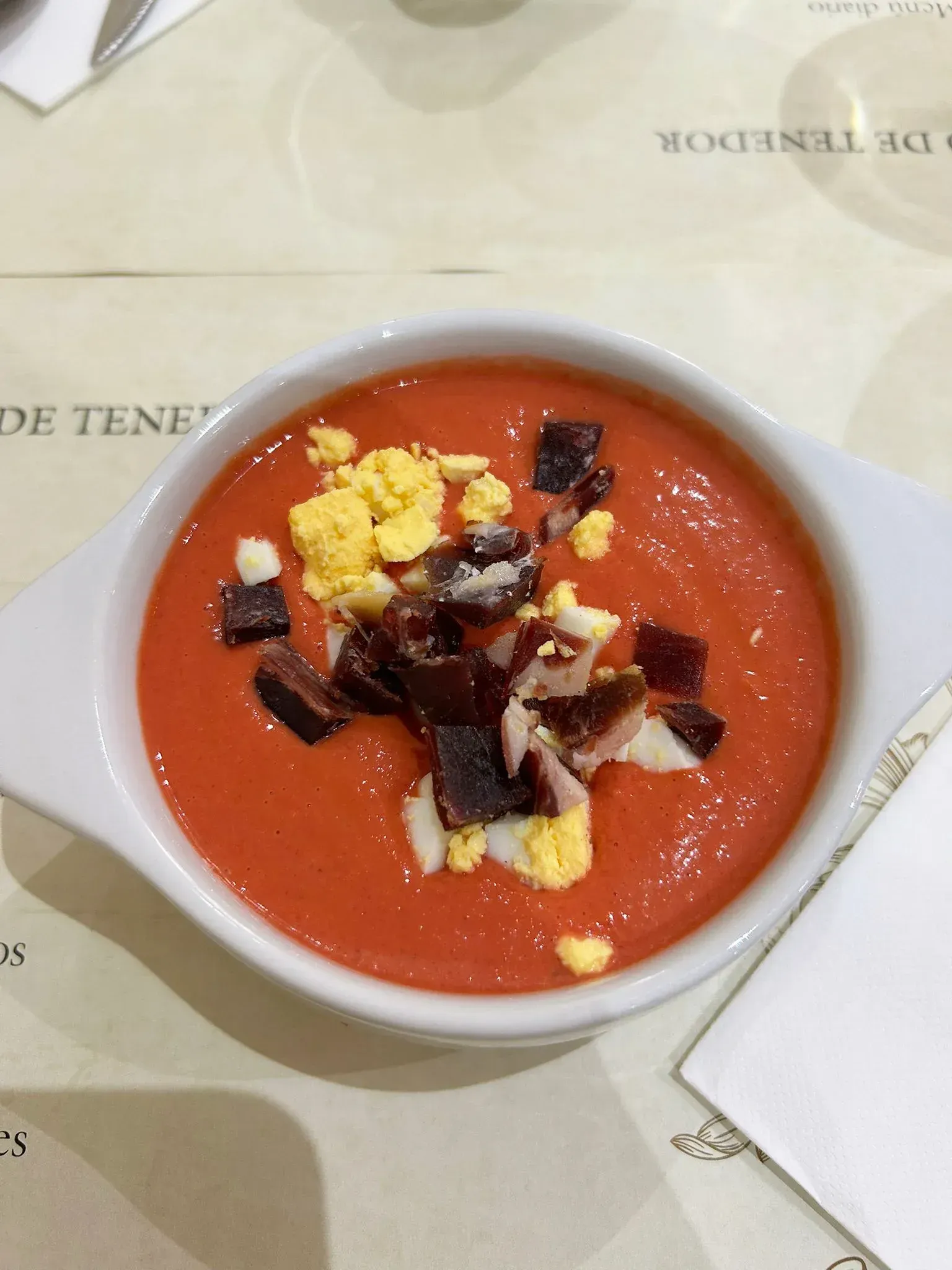 Sopa de gazpacho en recipiente blanco, cubierta con huevo picado y jamón, sobre la mesa.