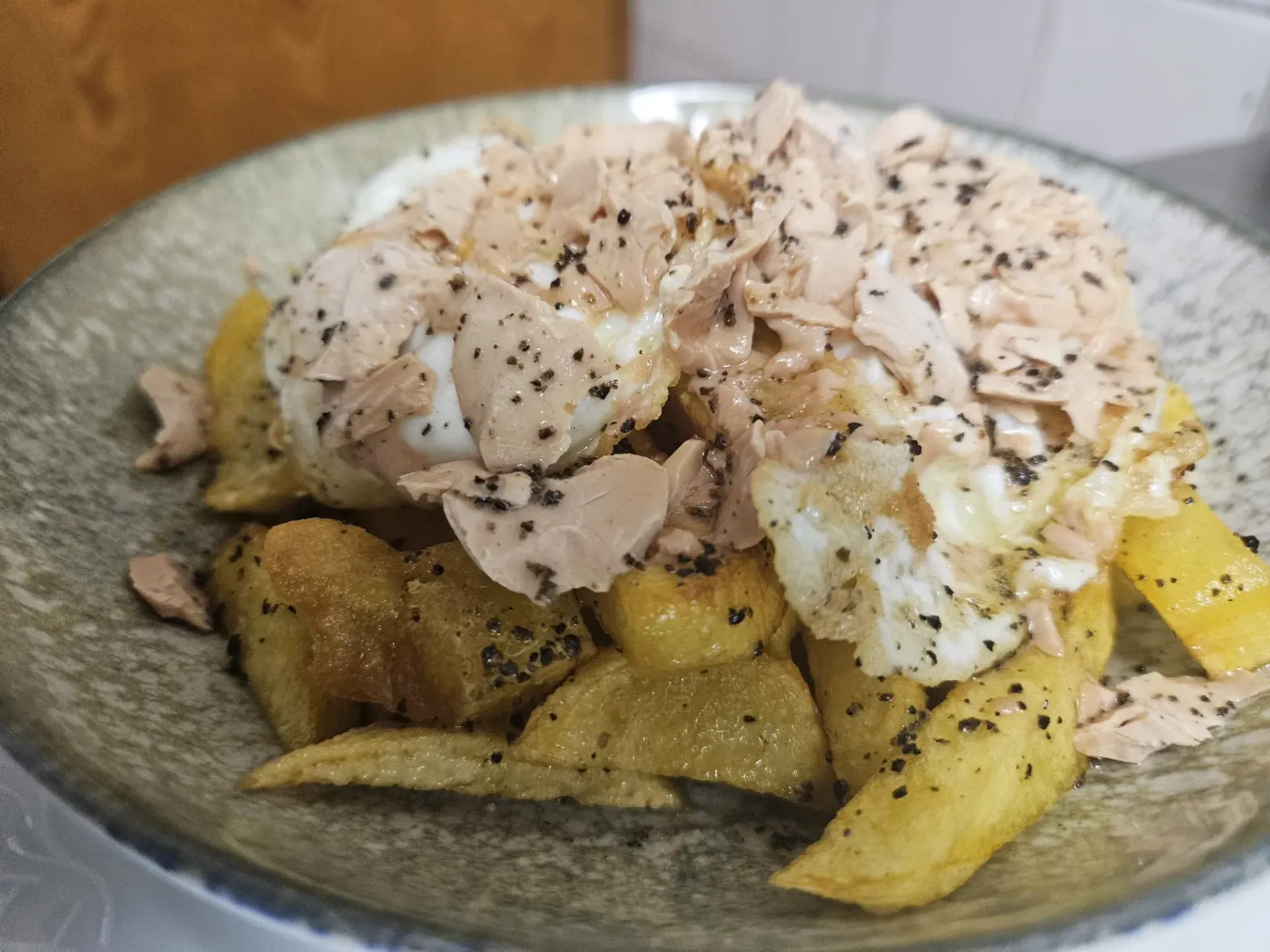 Huevos fritos y pescado desmenuzado sobre patatas fritas, espolvoreado con pimienta negra en un plato gris.