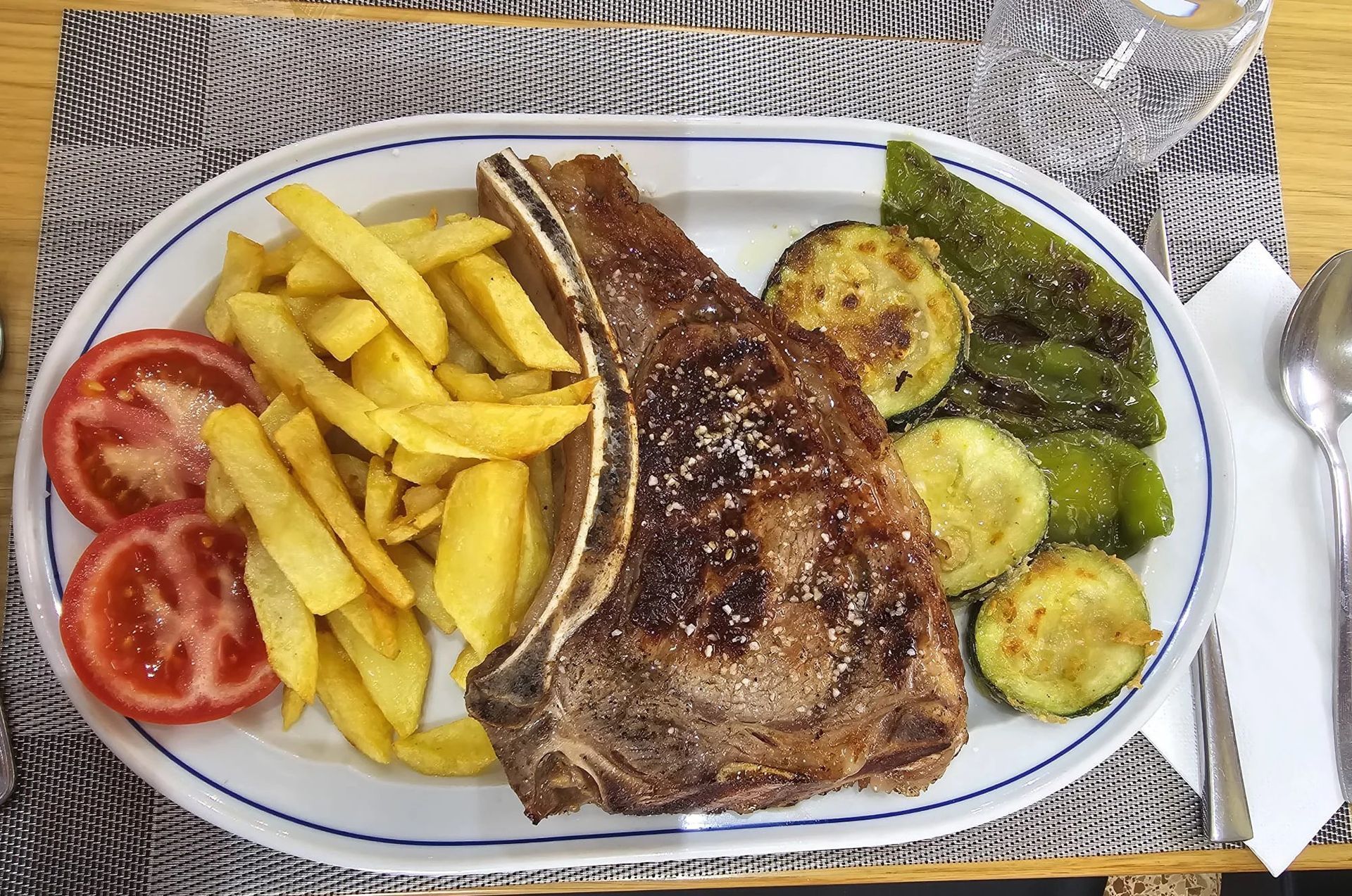 Bistec a la parrilla con patatas fritas, tomates y verduras a la parrilla en un plato blanco.