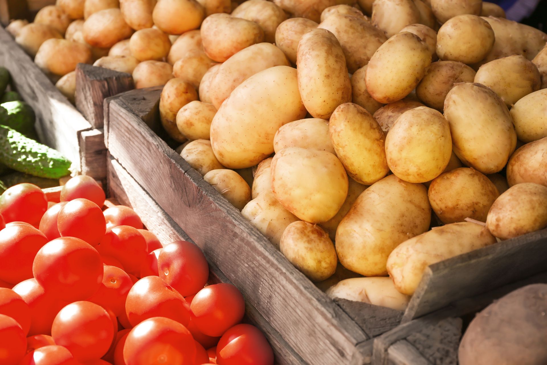 Tomates et pommes de terre dans des caisses en bois sur un marché, avec des légumes verts visibles.