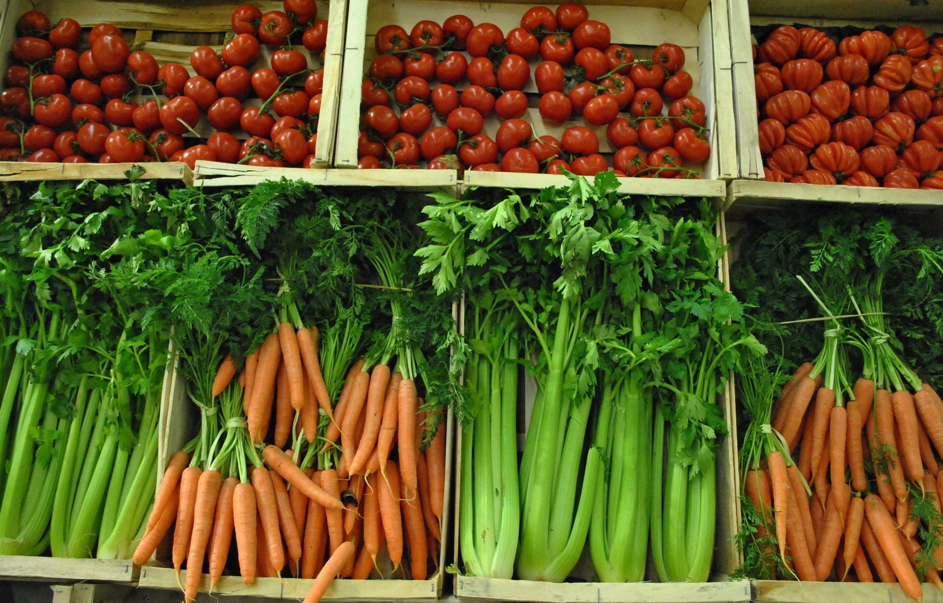 Des caisses en bois remplies de tomates rouges, de carottes et de céleris sont exposées sur un marché.