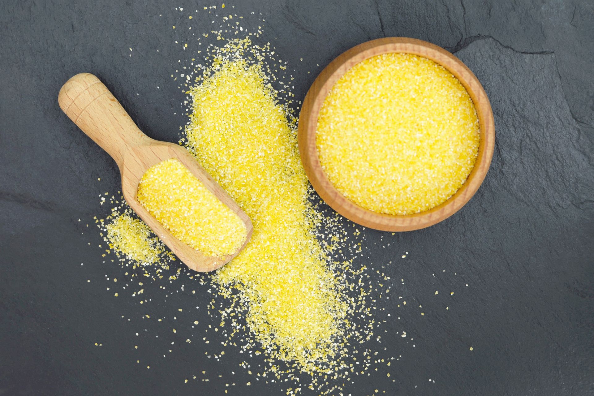 De la semoule de maïs jaune dans un bol en bois et une louche, se déversant sur une surface gris foncé.