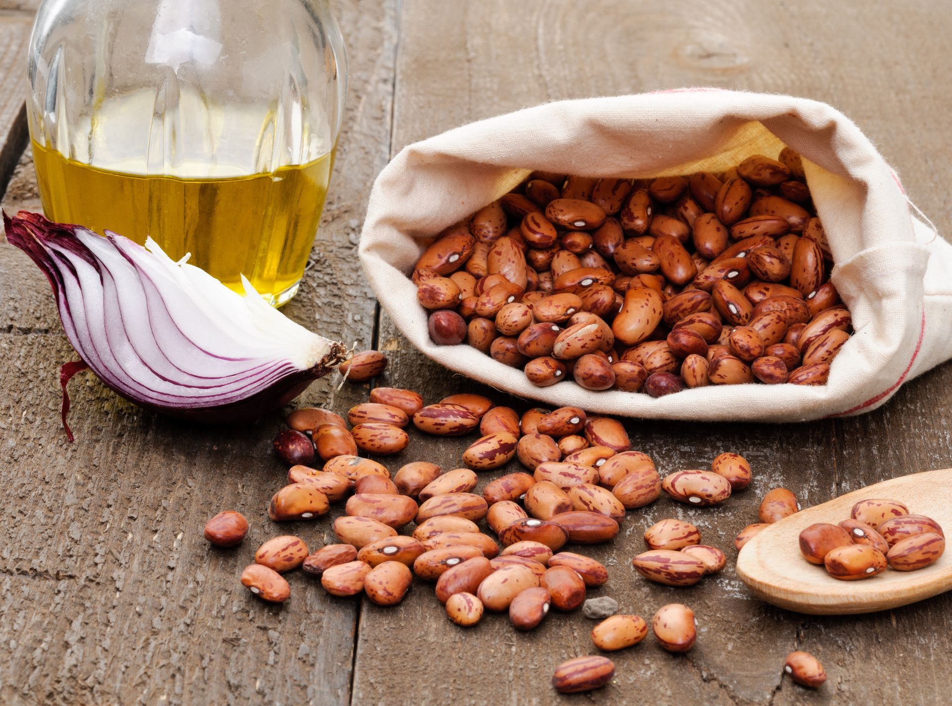 Des haricots débordent d'un sac en tissu, avec une tranche d'oignon rouge, de l'huile d'olive et une cuillère en bois sur une surface en bois.