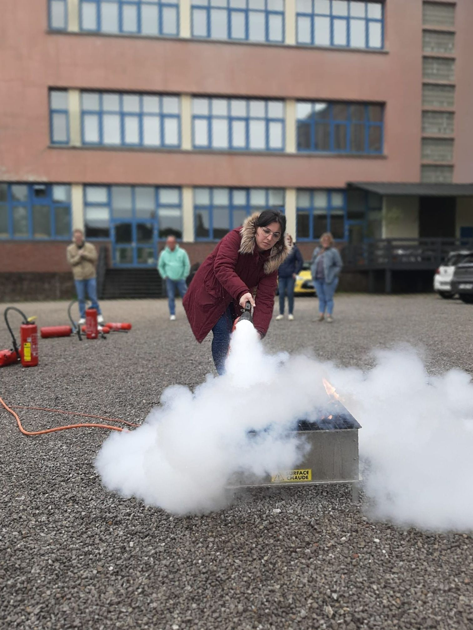 Une femme utilise un extincteur, créant un nuage de fumée blanche. Plusieurs personnes observent la scène en extérieur.