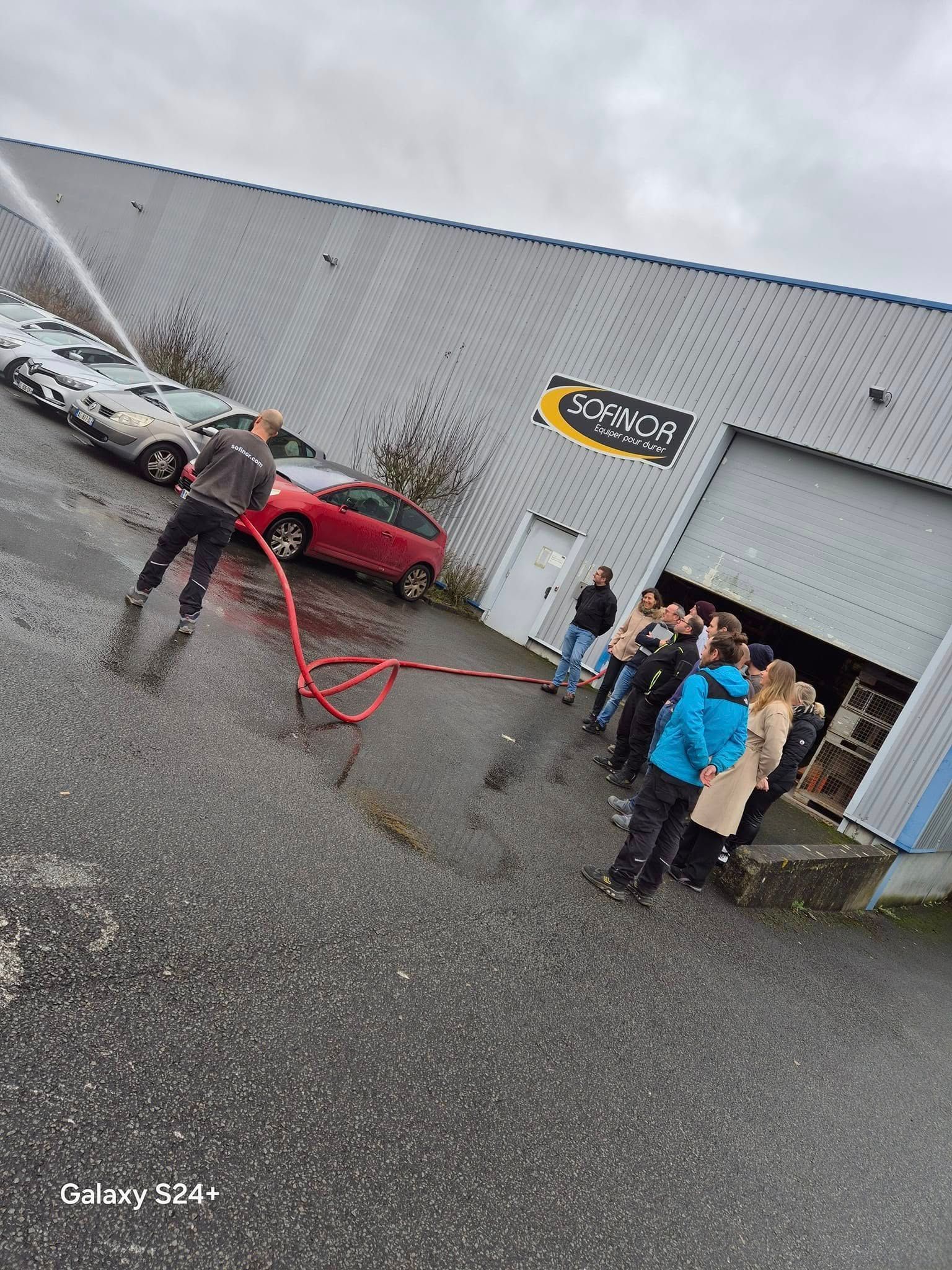 Une personne arrose avec un tuyau d'arrosage ; un groupe de personnes observe la scène depuis l'extérieur d'un bâtiment, des voitures se trouvant à proximité.
