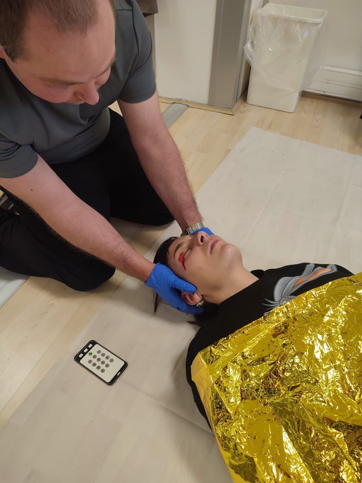 Une personne porte secours à un blessé allongé au sol, recouvert d'une couverture dorée.