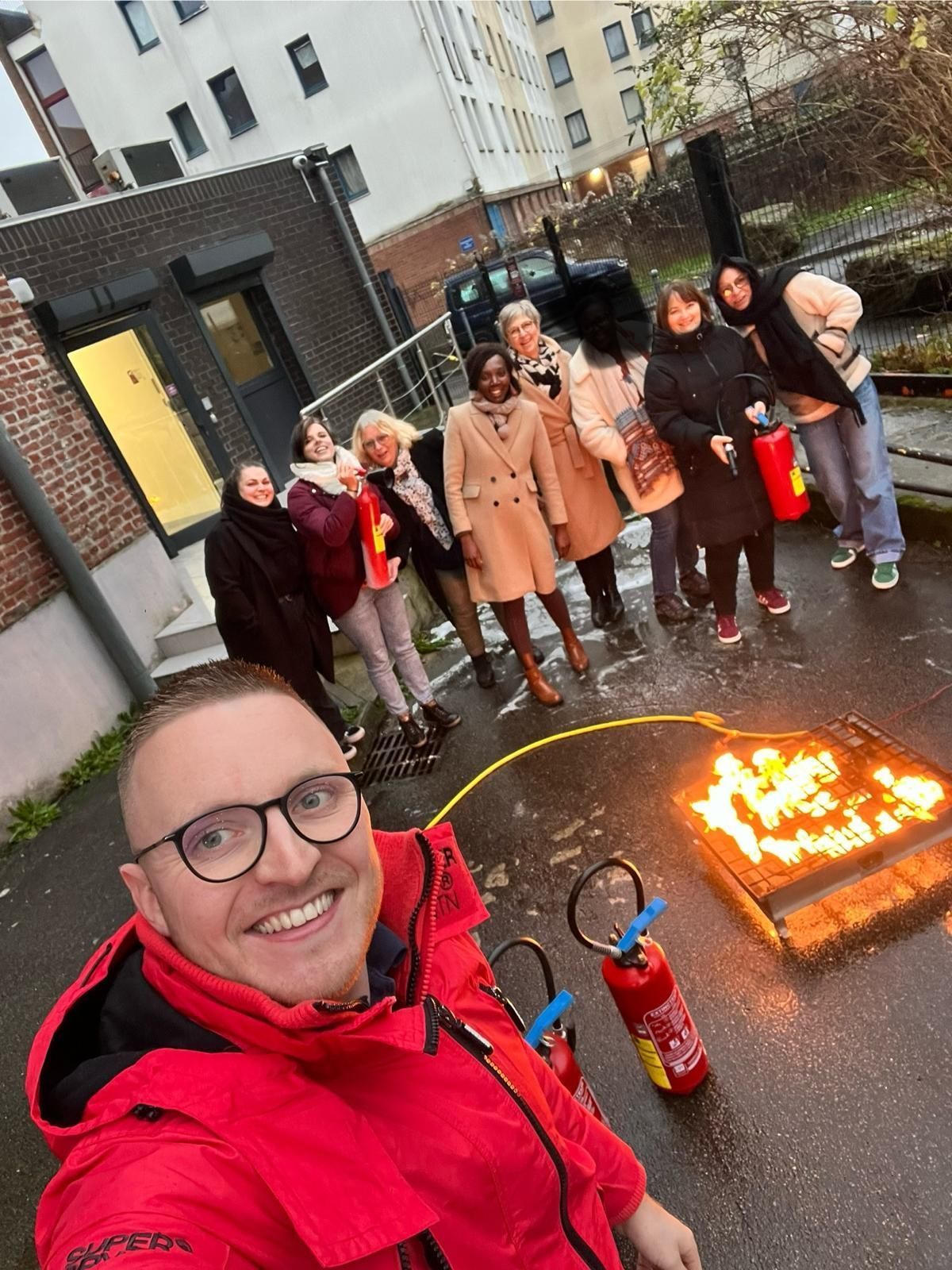 Un homme sourit, prend un selfie avec un groupe, un feu brûle dans une caisse ; des extincteurs se trouvent à proximité. Un bâtiment en briques apparaît en arrière-plan.