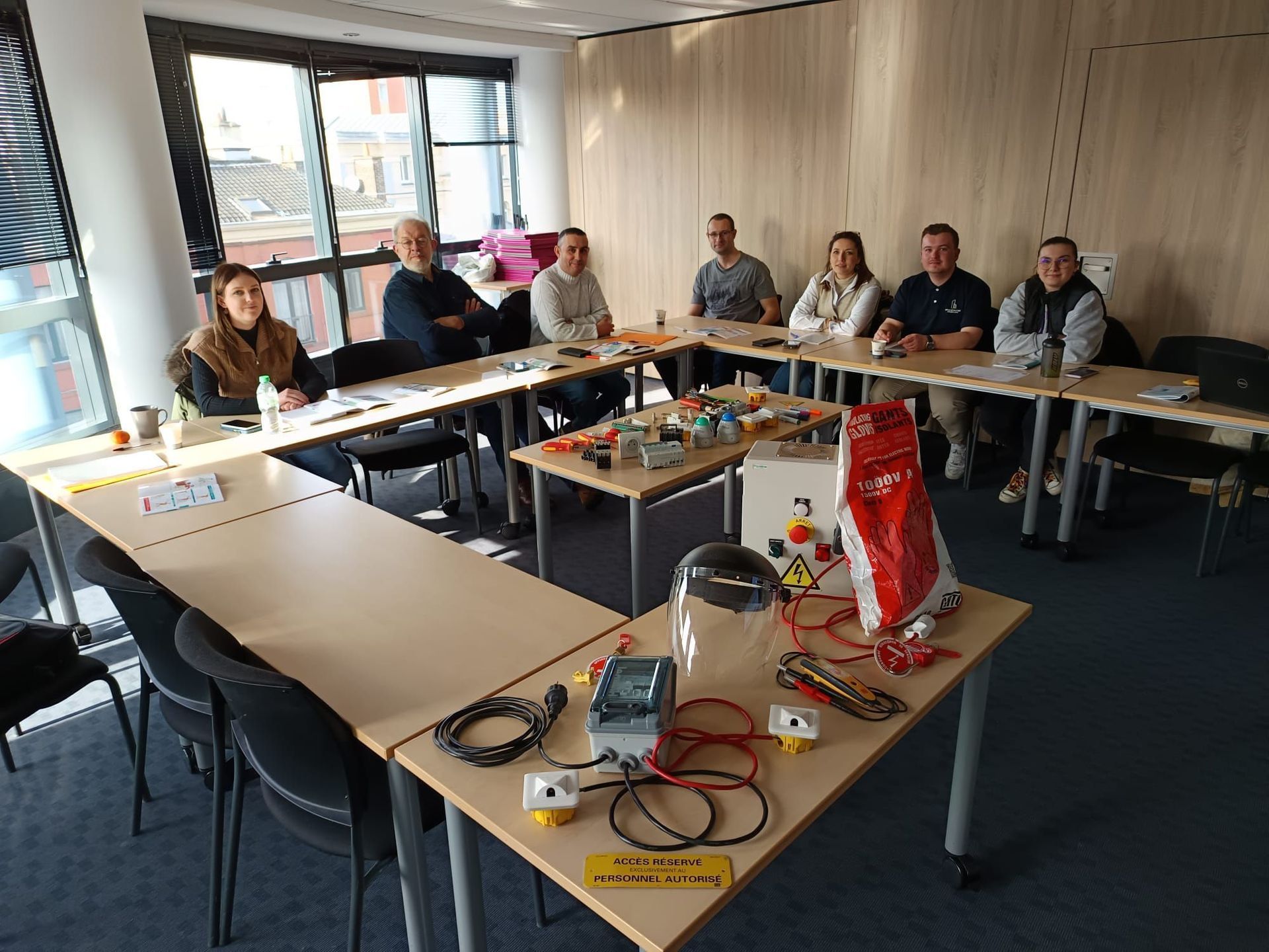 Des personnes assises autour de tables où sont exposés des appareils électriques, dans une pièce baignée de lumière.
