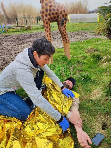 Une femme soigne une personne allongée au sol, la recouvrant d'une couverture dorée ; une statue de girafe se dresse en arrière-plan.