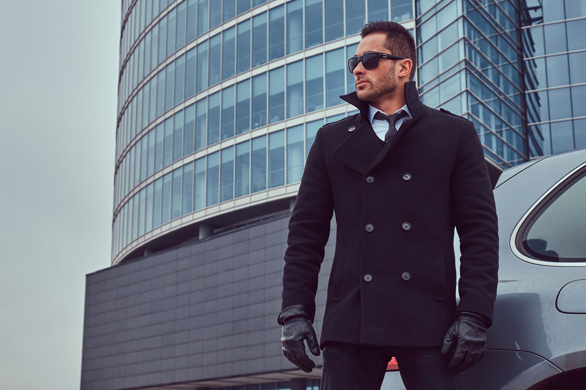 Un homme en manteau avec des lunettes de soleil se trouve à côté d'une voiture, un bâtiment moderne se dresse en arrière-plan.