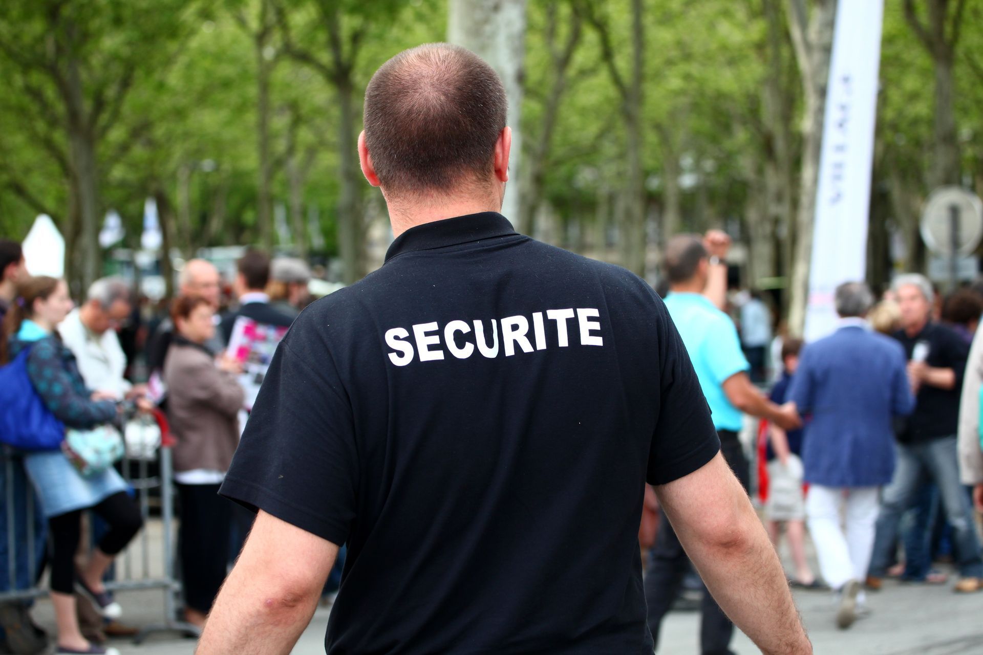 Agent de sécurité en chemise noire avec l'inscription SÉCURITÉ dans le dos, observant la foule. En extérieur.