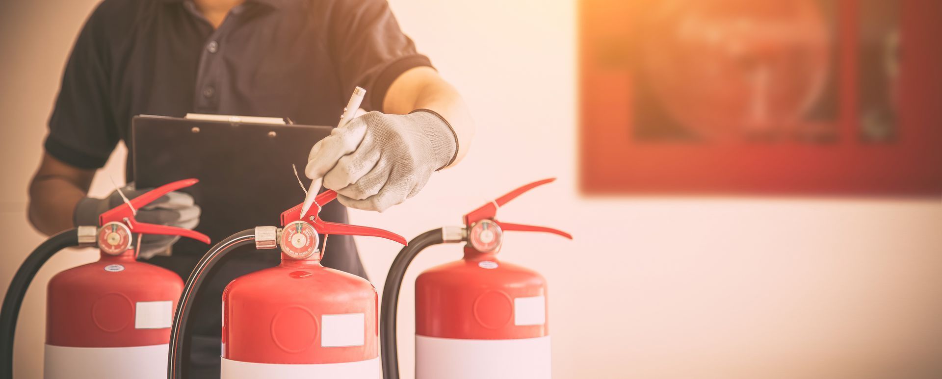 Une personne vêtue d'une chemise sombre et portant des gants inspecte des extincteurs, tenant un bloc-notes devant un mur.