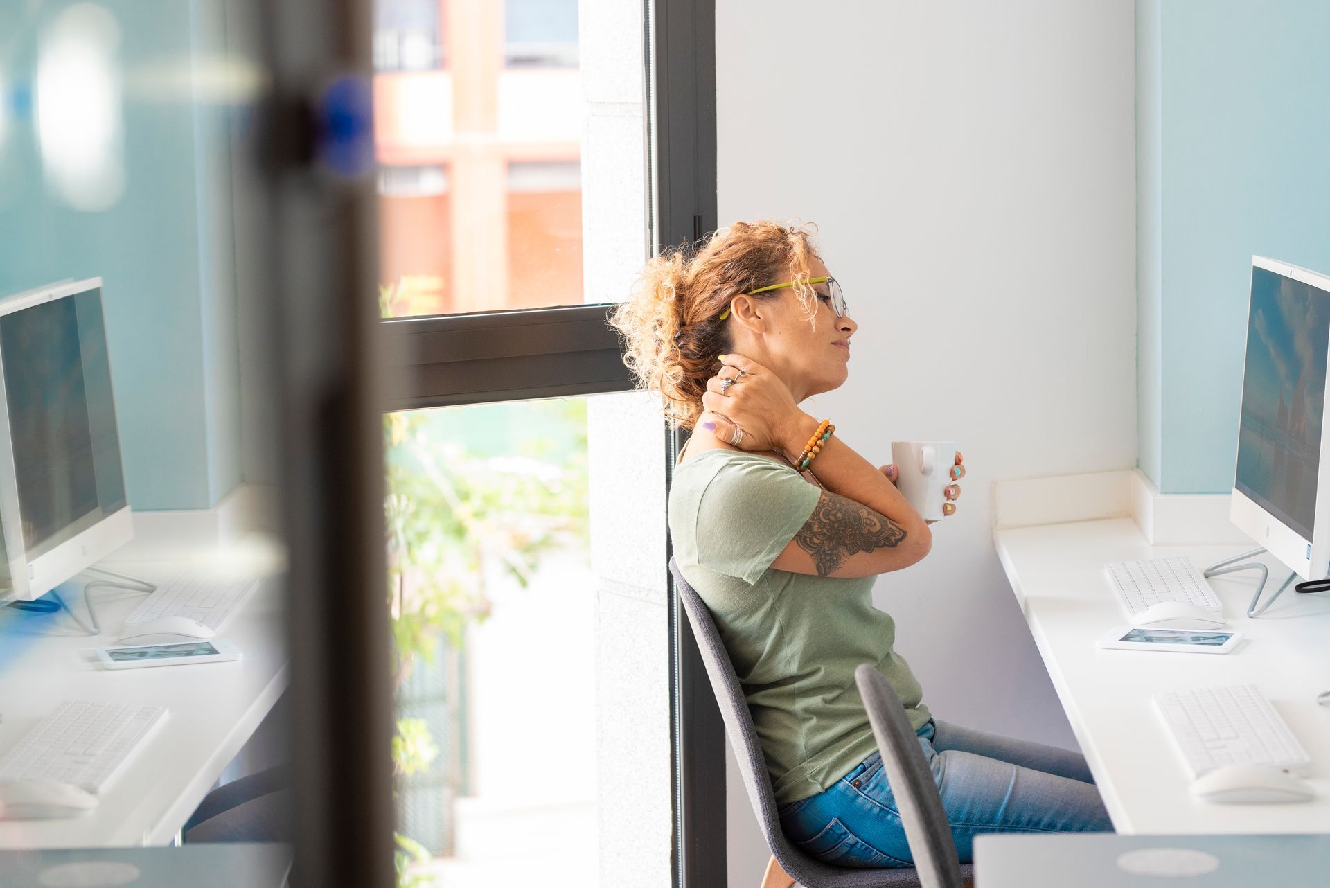 Femme assise à un bureau, tenant une tasse, se touchant la nuque, détournant le regard de son ordinateur, bureau vitré.