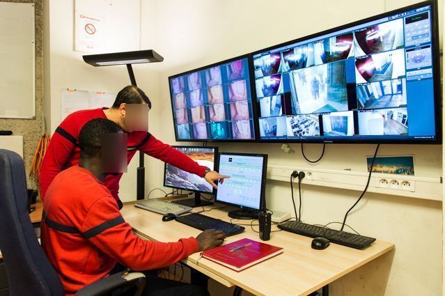 Dans une salle de sécurité, deux personnes surveillent plusieurs écrans affichant des images de vidéosurveillance. L'une d'elles désigne un ordinateur.