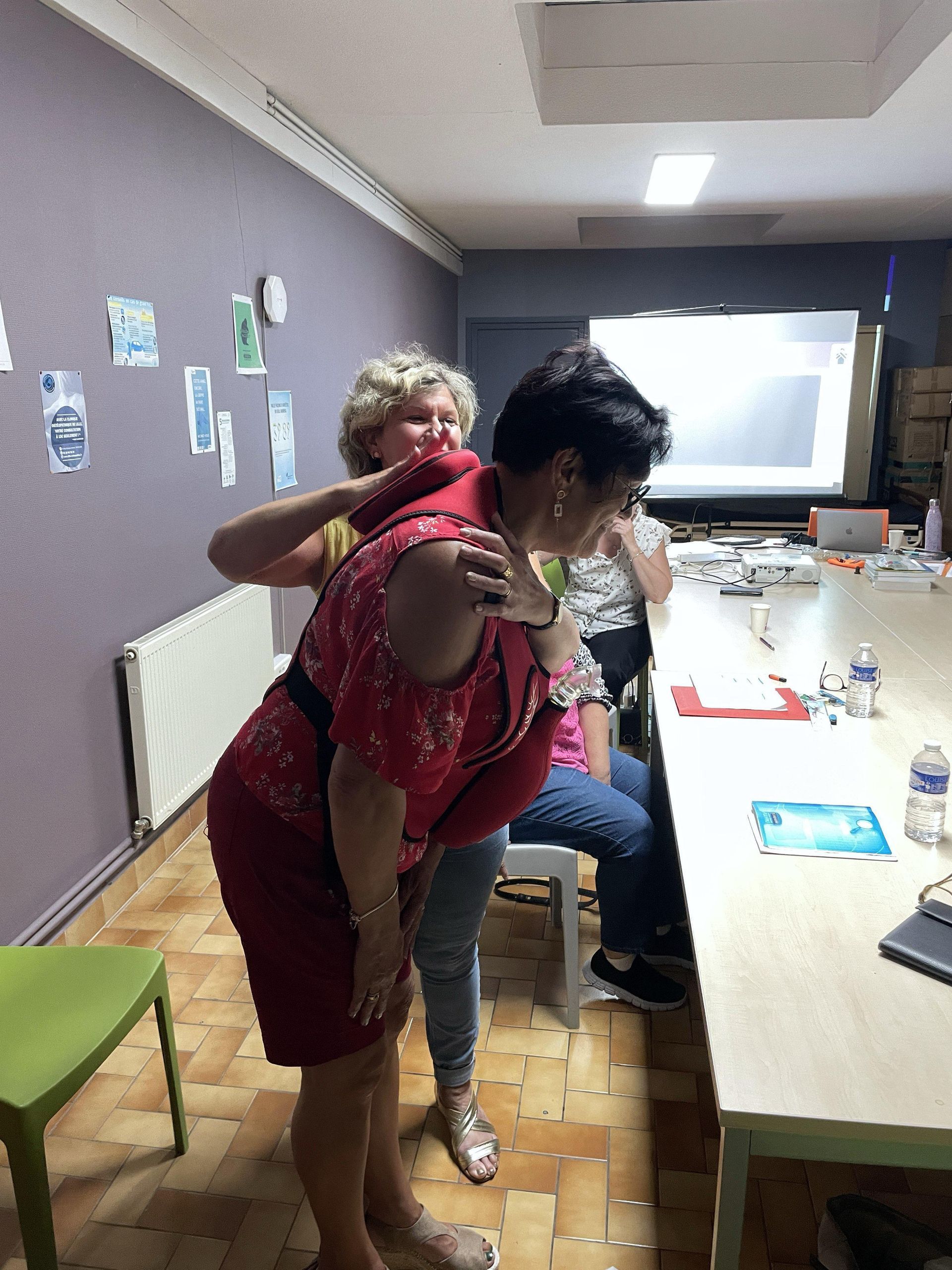 Une femme en robe rouge est aidée par une autre femme assise à une table.