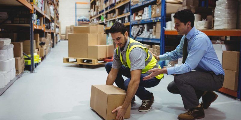 Un employé dans un entrepôt soulève un carton, sous la supervision et les gestes d'un responsable.
