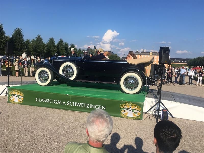 Ein Mann steht vor einem schwarzen Auto auf einer grünen Bühne.