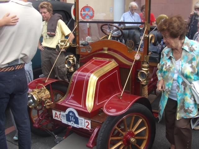 Eine Gruppe von Menschen schaut sich ein altes rotes Auto mit einem Nummernschild an, auf dem steht: ac