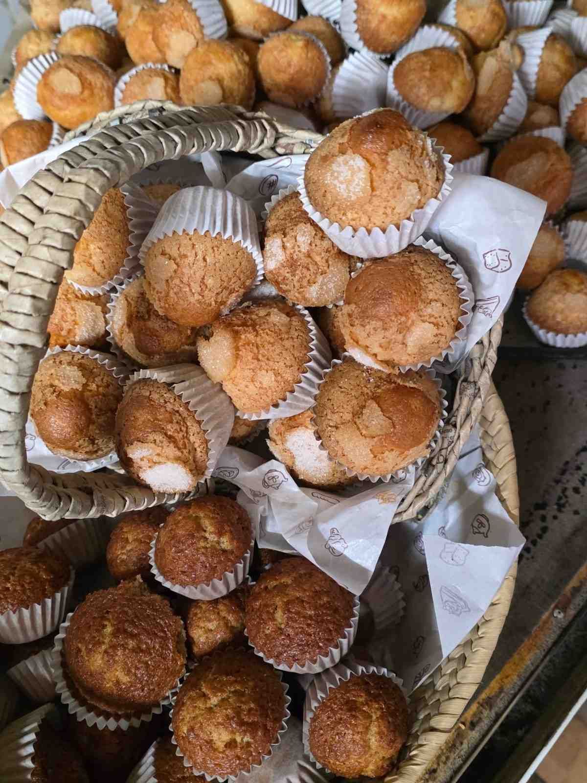 Una cesta de mimbre rebosante de magdalenas doradas en moldes de papel blanco, rodeada de más magdalenas sobre una encimera.