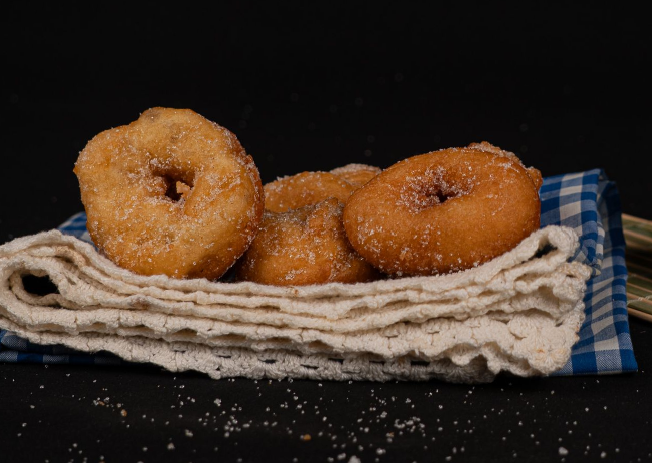 Tres rosquillas de canela sobre un paño junto a una taza gris con una cuchara sobre un fondo negro.