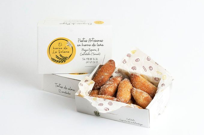 Caja de panadería blanca con pasteles y un paquete con etiqueta dorada sobre un fondo blanco brillante.