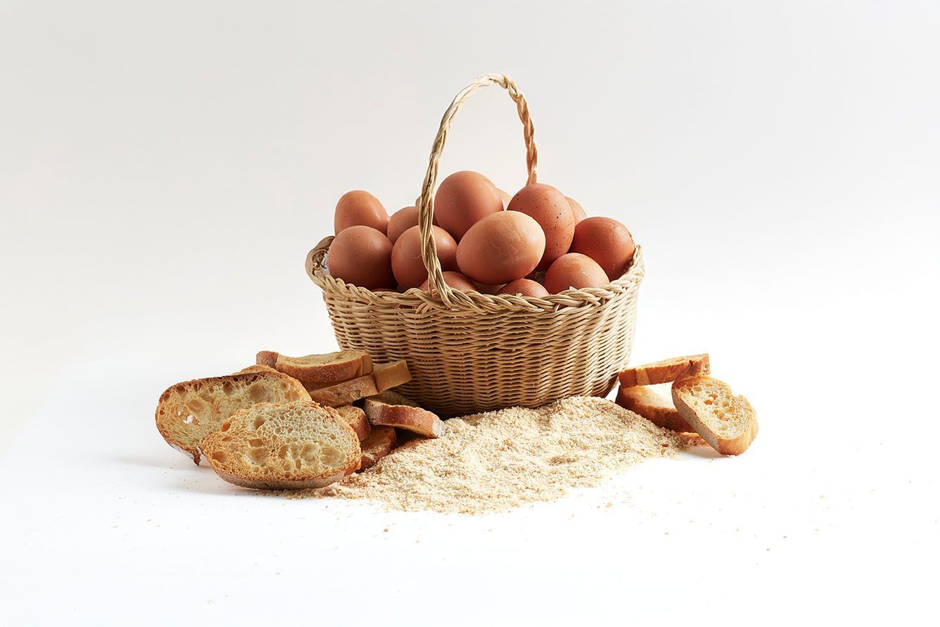 Cesta de mimbre llena de huevos junto a panes sobre un fondo blanco.
