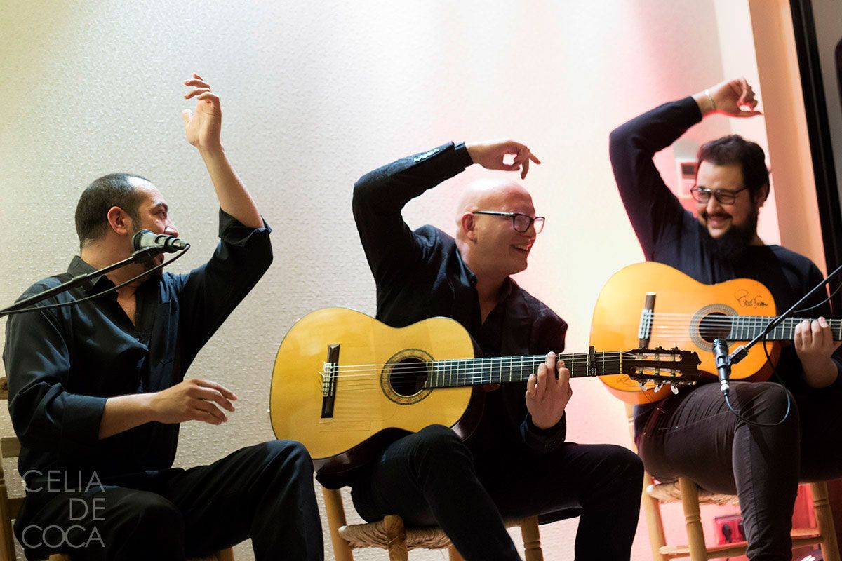 Tres hombres tocan guitarras y cantan con micrófonos frente a un cartel que dice Celia de Coca.