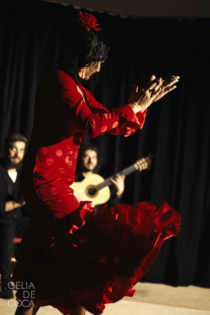 Una mujer con un vestido rojo baila flamenco con un hombre tocando una guitarra en el fondo.