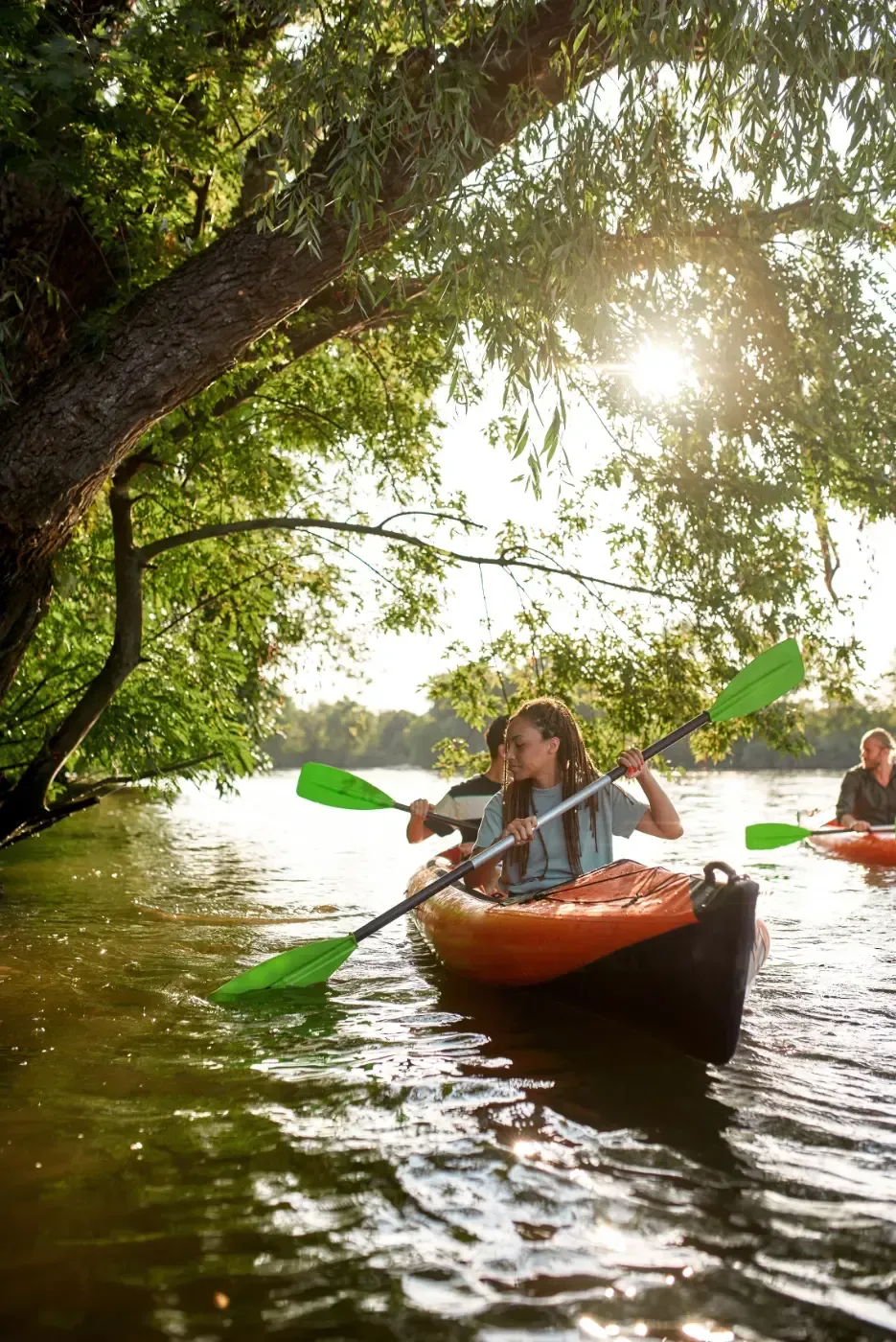 Zwei Personen paddeln in einem orangefarbenen Kajak auf einem sonnenbeschienenen Fluss unter den Ästen eines großen Baumes.