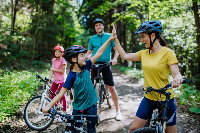 Eine Familie mit Helmen fährt mit Fahrrädern auf einem Waldweg; im Vordergrund geben sich ein Kind und ein Erwachsener ein High-Five.