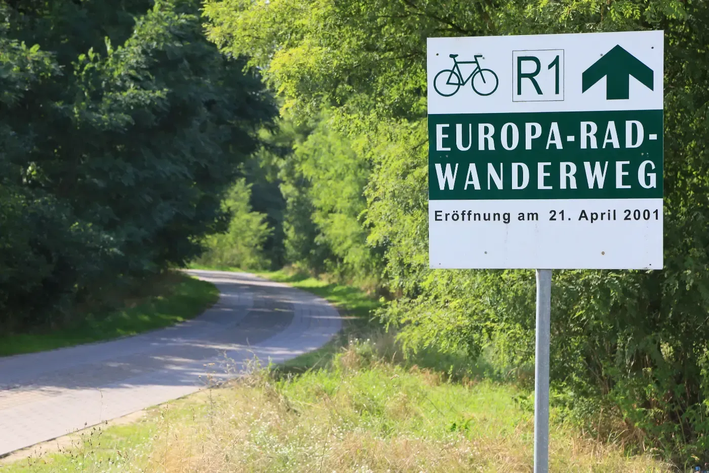 Ein Schild mit der Aufschrift „Radweg R1 Europa-Rad-Wanderweg“ steht auf einem Pfosten neben einem ländlichen, gepflasterten Weg, der von Bäumen gesäumt ist.