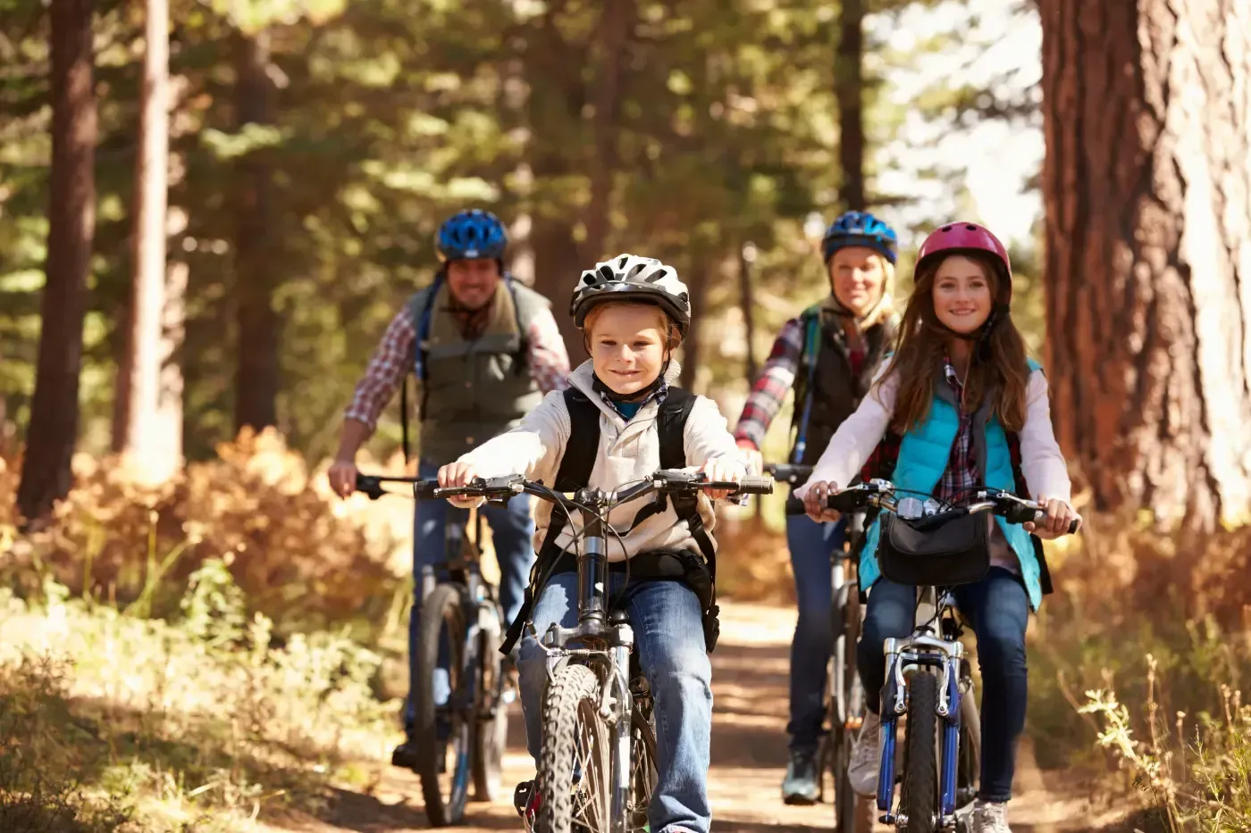 Eine Familie mit Helmen fährt mit Mountainbikes auf einem sonnenbeschienenen Feldweg durch einen Wald.