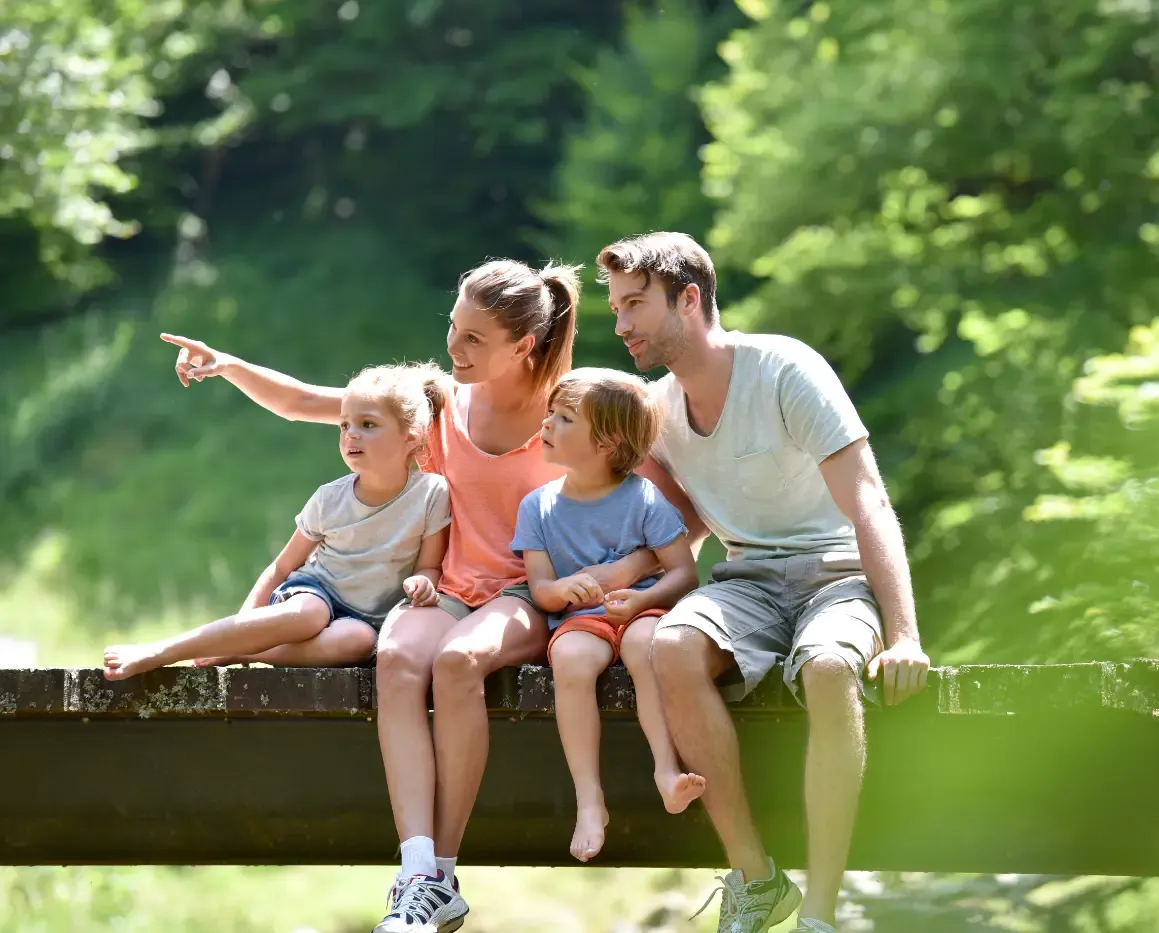 Eine vierköpfige Familie sitzt auf einer Holzbrücke in einem sonnenbeschienenen Wald, wobei die Mutter in die Ferne zeigt.
