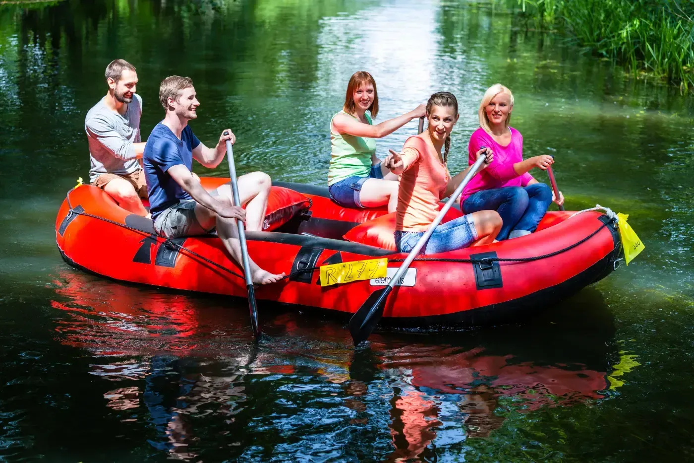 Vier lächelnde Menschen paddeln auf einem leuchtend roten Schlauchboot einen ruhigen, sonnenbeschienenen Fluss hinunter, der von viel Grün umgeben ist.