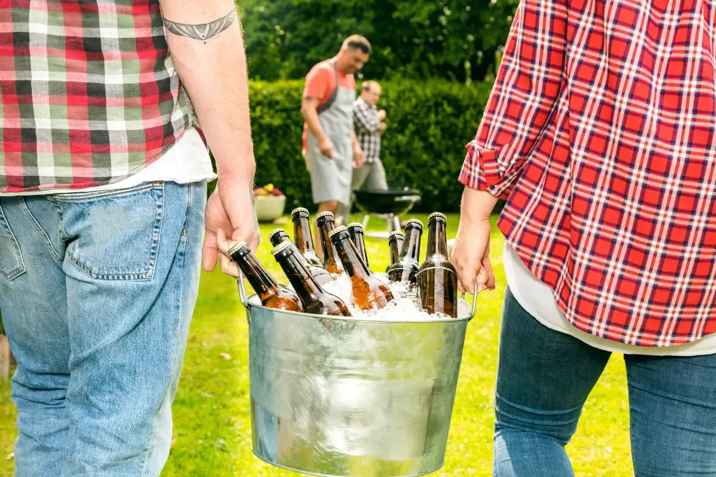 Zwei Personen in karierten Hemden gehen über einen Rasen und tragen einen Metalleimer, der mit Eis und Glasflaschen gefüllt ist.