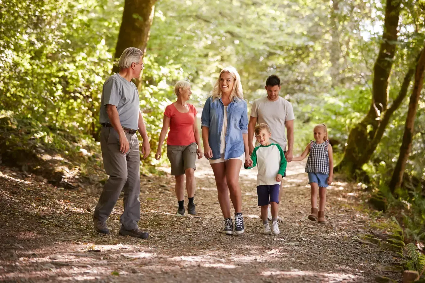 Eine Familie mehrerer Generationen spaziert gemeinsam auf einem sonnenbeschienenen, von Bäumen gesäumten Feldweg in einem Wald.