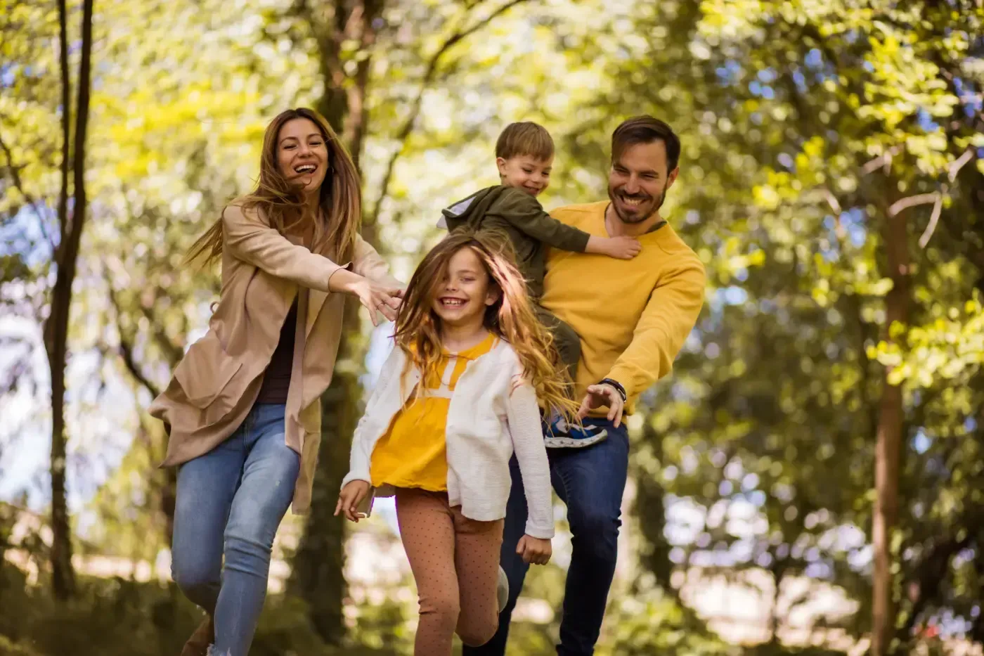 Eine fröhliche Familie rennt gemeinsam durch einen sonnendurchfluteten, bewaldeten Park und lacht und lächelt bei einer Aktivität im Freien.
