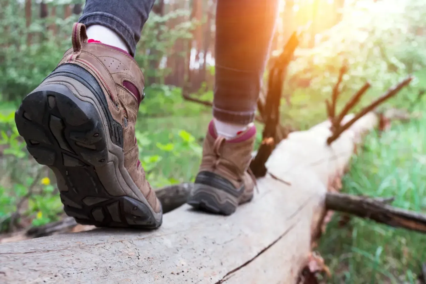 Nahaufnahme einer Person in braunen Wanderschuhen, die auf einem umgestürzten Baumstamm in einem Wald entlanggeht.