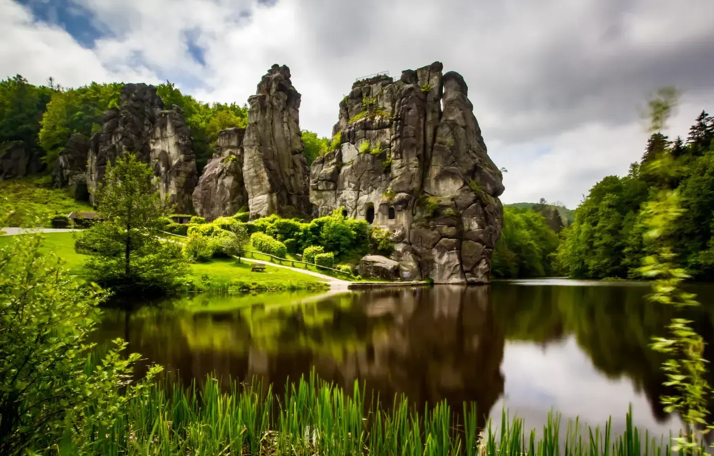 Die Externsteine ​​erheben sich über einem ruhigen Teich, umgeben von üppig grünen Bäumen unter einem bewölkten Himmel.