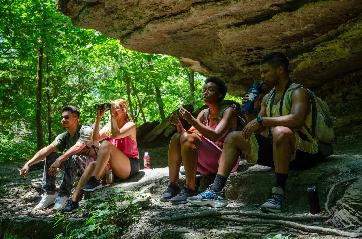 Vier Personen sitzen unter einem Felsvorsprung in einem üppigen, grünen Wald und machen eine Pause während ihrer Wanderung.